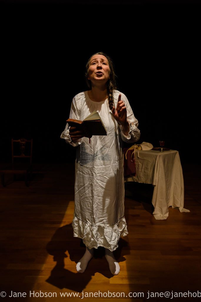 A full length photograph of a white actress in a white gown standing with a book in her hand, reading from it to the audience