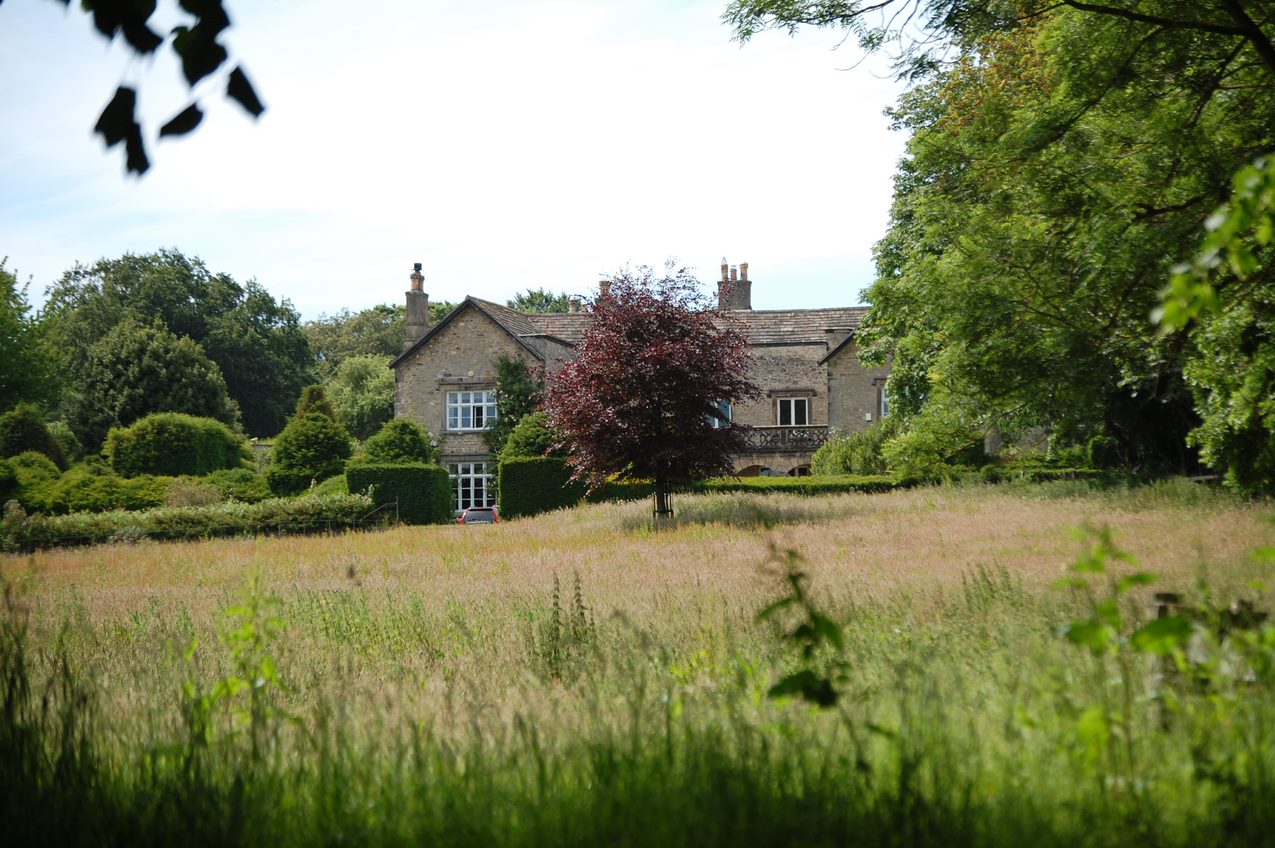 An old house of grey stone sits in the background of a verdant lawn, with trees and long grass.